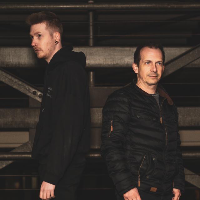 Zwei Männer stehen Rücken an Rücken in dunkler Kleidung vor Metallstruktur