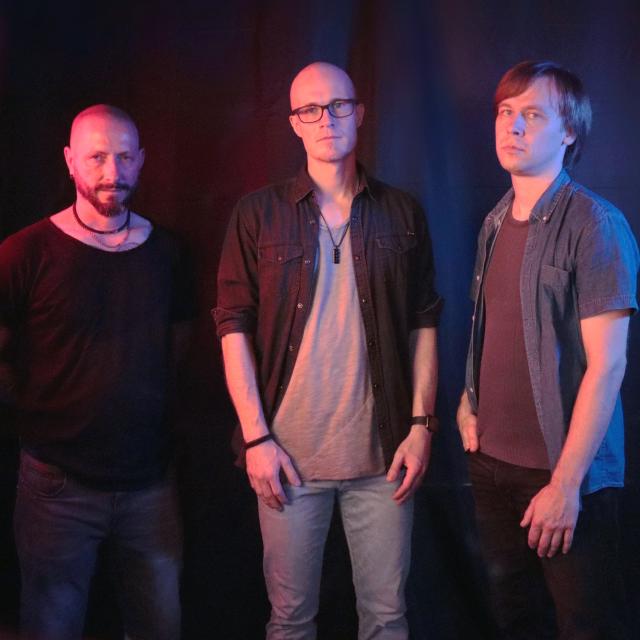 Drei Personen stehen nebeneinander vor dunklem Hintergrund, lässig gekleidet, leicht farbig beleuchtet.