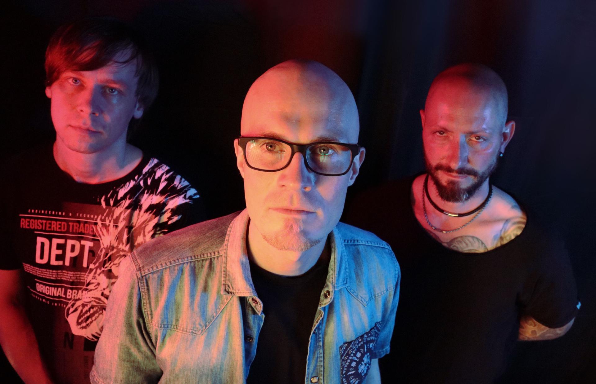 Drei Männer stehen nebeneinander vor dunklem Hintergrund, farbig beleuchtet und schauen zur Kamera.