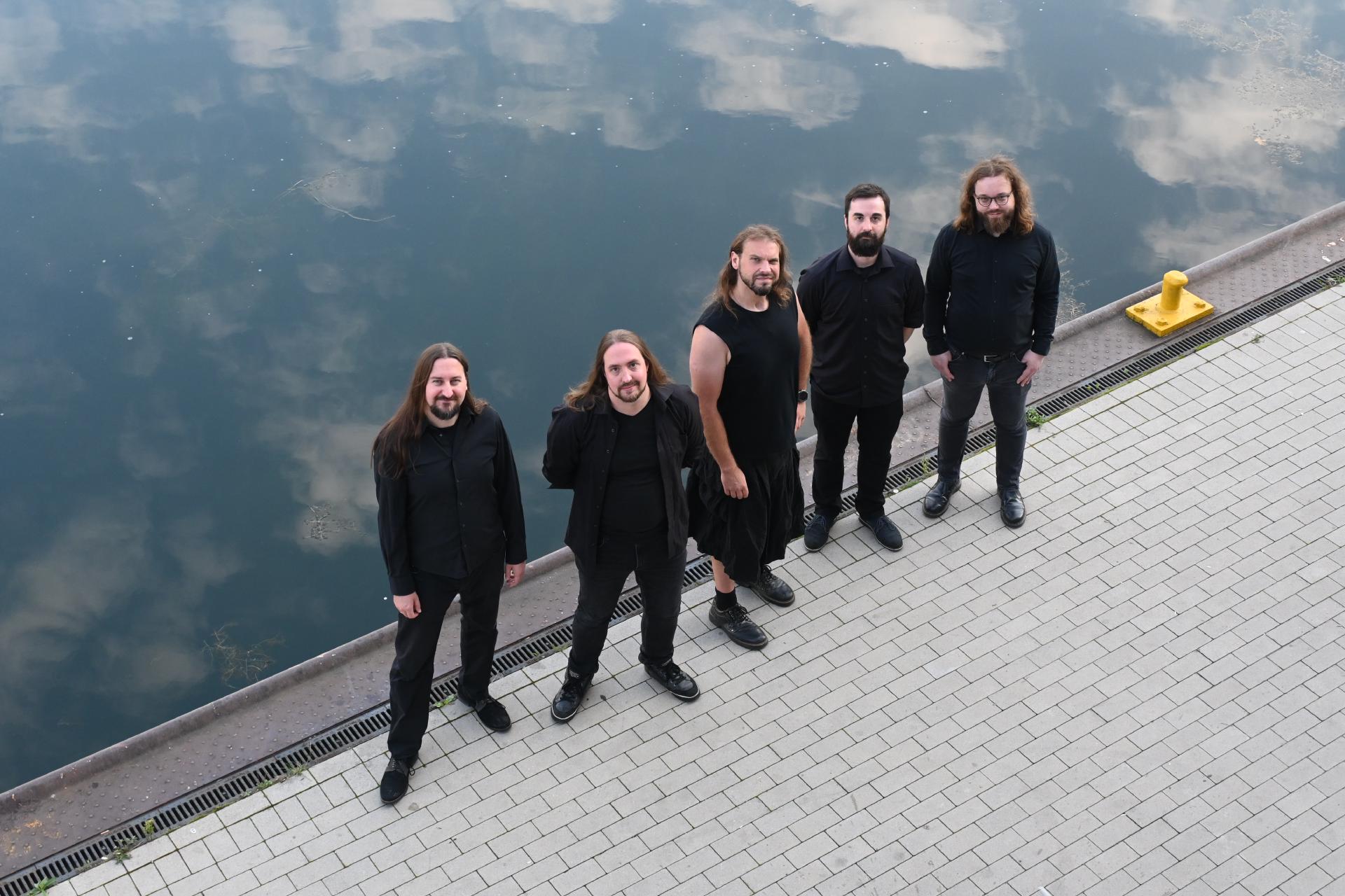 Die Band Oceansoul steht auf einer Kaimauer an einem ruhigen, trüben Gewässer.