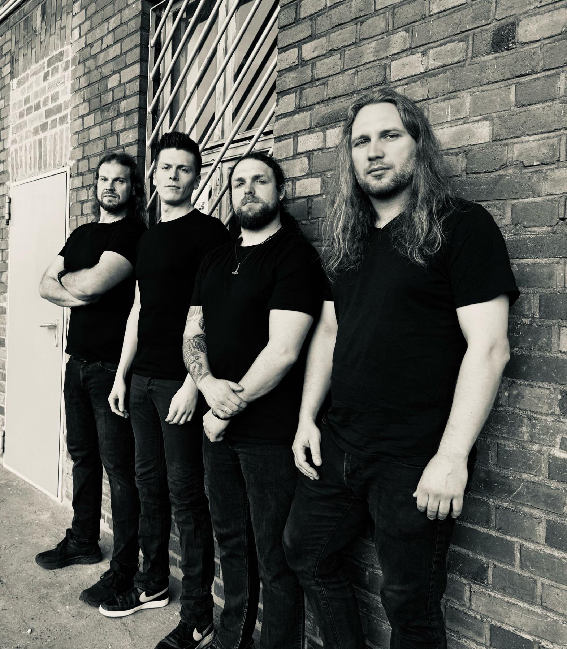 Vier gut aussehende, mehr oder weniger junge Männer. Drei mit langen Haaren. Schwarz-Weiß vor einer industriellen Hausfassade.