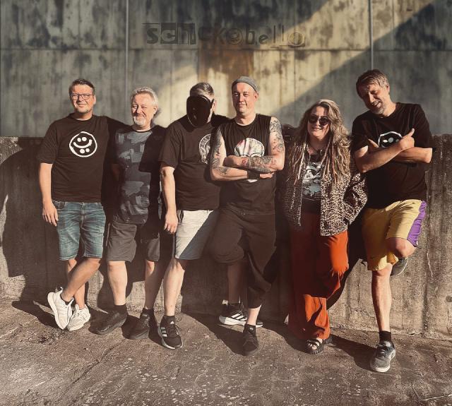 Sechs Personen posieren fröhlich nebeneinander an einer Betonwand, sommerlich gekleidet bei Sonnenlicht.