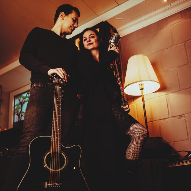 Zwei Personen stehen nah beisammen, warmes Licht, Gitarre und Mikrofon in den Händen.